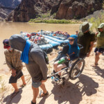 Two people carrying a woman in a wheelchair.